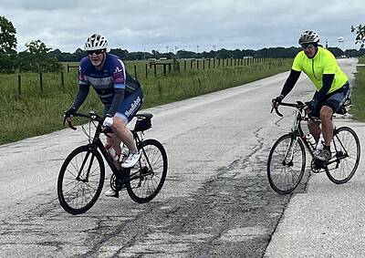 Two cyclists riding on a rural road during daytime, promoting Bluebonnet Express bike tours and cycling events.