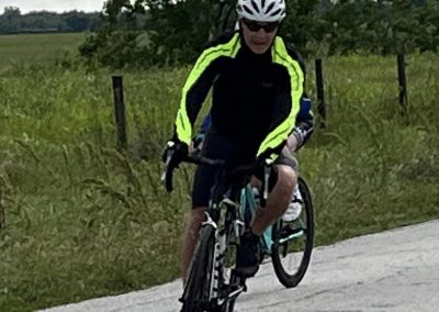 Bright cyclist riding a bike on rural road with green fields and cloudy sky in the background.