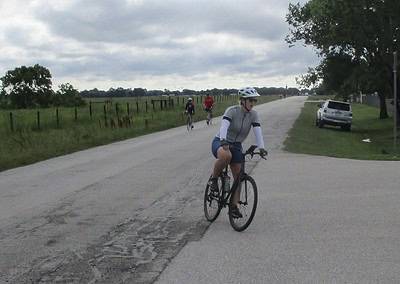 Cyclist riding on a rural road with others biking in the background under cloudy skies.