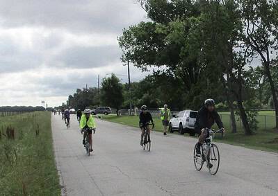 Bike riders on scenic rural road with trees and cloudy sky.
