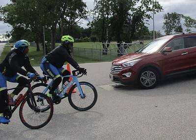 Cyclists riding bikes across a street with a red vehicle passing by, part of Bluebonnet Express bike route.