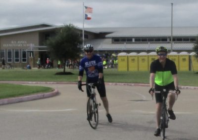 Runners in cycling event at Bluebonnet Express, Texas, with spectators and school building in background.