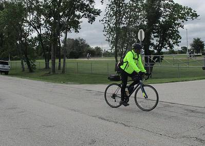 Cyclist in bright green jacket riding past a park with trees and a fence.