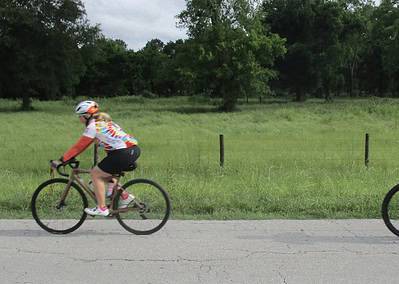 Brightly colored cyclists riding on a scenic bike trail surrounded by lush green trees and open grass fields.