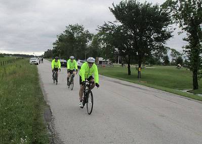 Bright yellow cycling jackets on group of cyclists riding on rural road during overcast day.