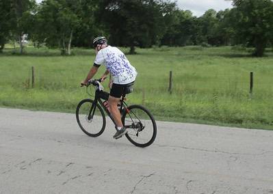 Vibrant cyclist riding a bike along a rural road with green fields and trees, promoting Bluebonnet Express cycling adventure and outdoor activity.