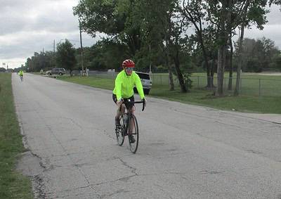 Bright yellow cycling jacket, cyclist riding on a paved road during daytime in overcast weather, Green trees in background.