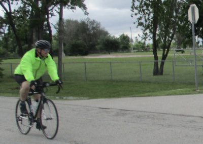 Bright green cyclist riding a road bike along a park trail with trees and cloudy sky in the background.