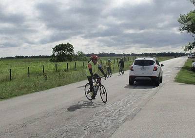 Cyclists riding on the road near grassy fields with cars parked alongside, promoting eco-friendly transportation.