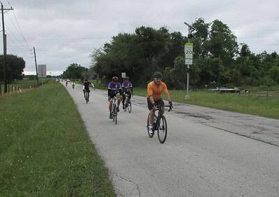 Cyclists riding on a rural road during a group ride with trees and overcast sky in the background.