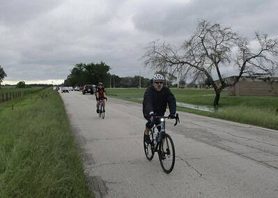 Brighton cycling on rural road in cloudy weather with green grass and trees in background.