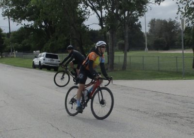 Cyclists riding through a park on a sunny day with trees and a grassy area in the background.