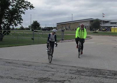 Two cyclists riding on a paved path outside a school with overcast skies, promoting Bluebonnet Express cycling event.