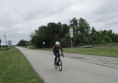 Cyclist riding on open road with greenery, safety helmet, and overcast sky for Bluebonnet Express bike route.