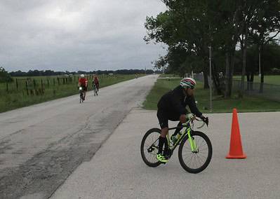 Cyclist riding near orange safety cone on rural bike trail with lush greenery and overcast sky.