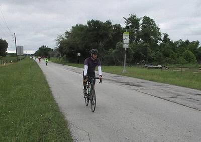 Cyclist riding on a rural road with greenery and overcast sky, part of Bluebonnet Express bike route.