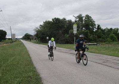 Outdoor cyclists riding on a rural road with greenery and overcast skies.