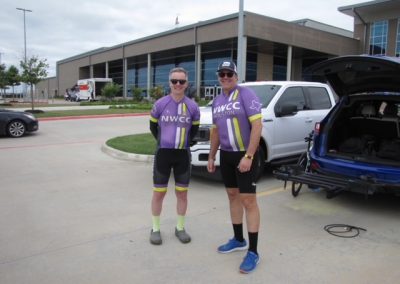 Two cyclists in NWCC jerseys standing outside in parking lot near support vehicle before Bluebonnet Express ride.