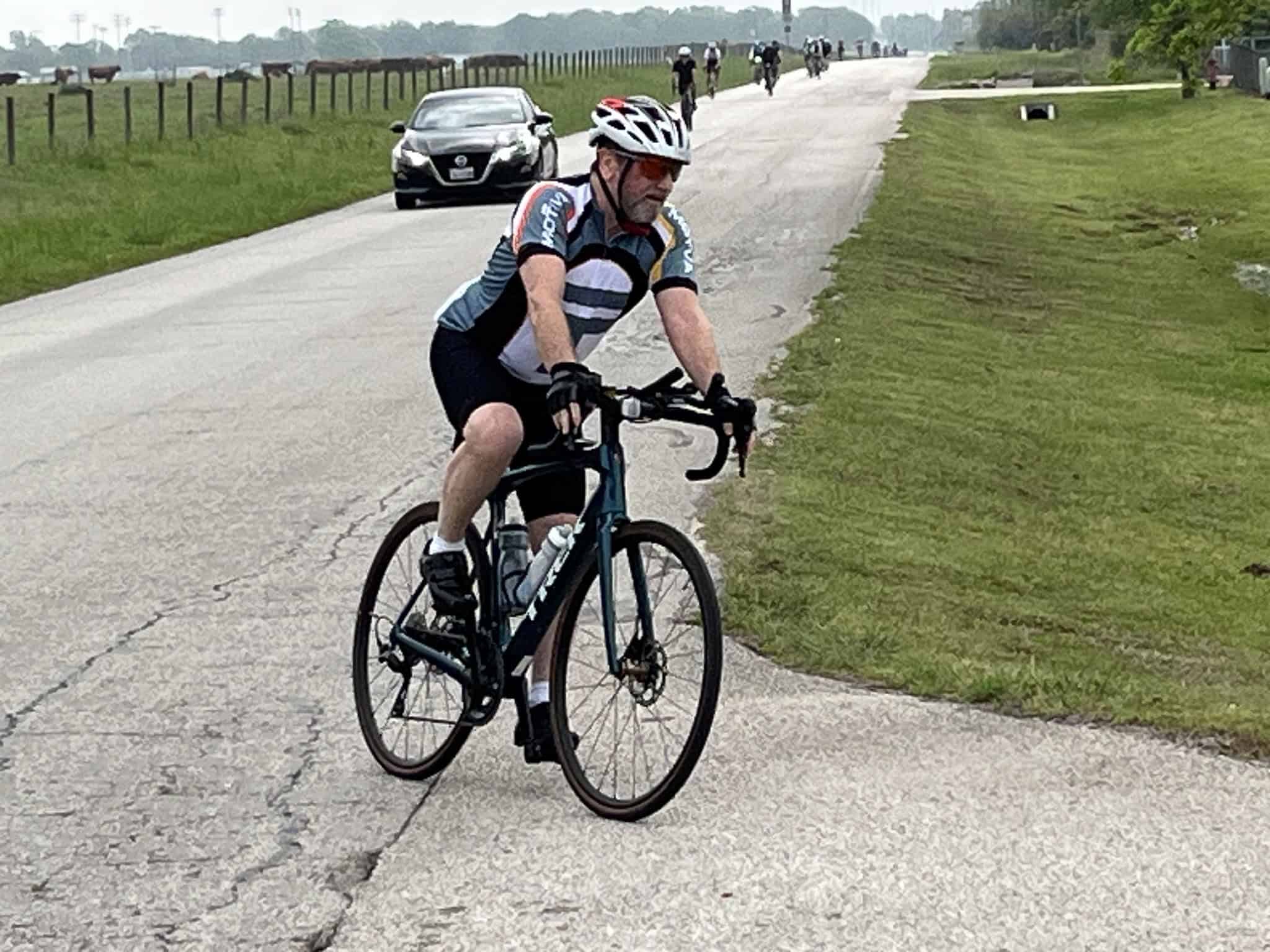 Cyclist riding on grassy shoulder with rural road, cars, and other riders in background.