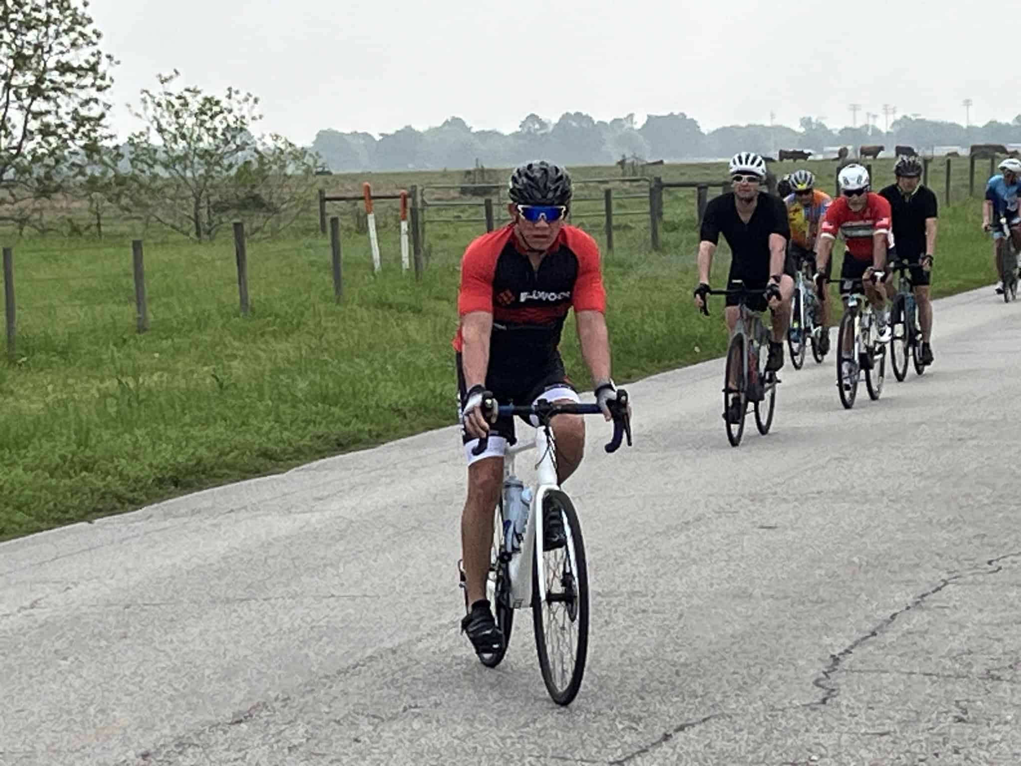 Velo riders cycling on rural road in Texas with green fields and horses in background.