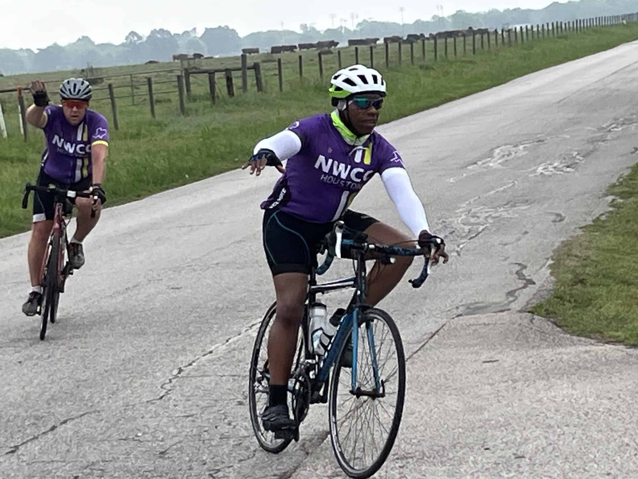 Mid-distance cyclists wearing NWCC Houston jerseys riding on a rural road, eco-friendly outdoor biking event.