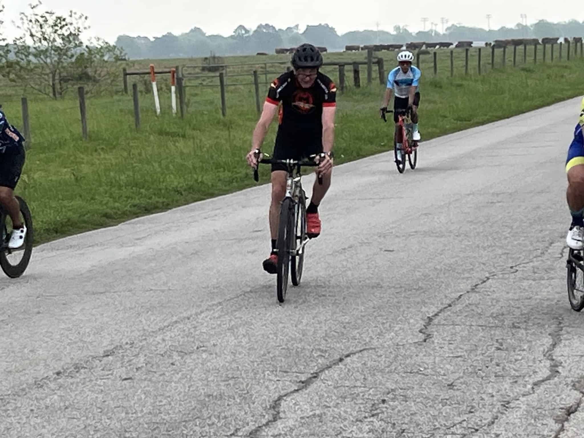 Cyclists riding on a scenic countryside road with lush green fields and fencing.
