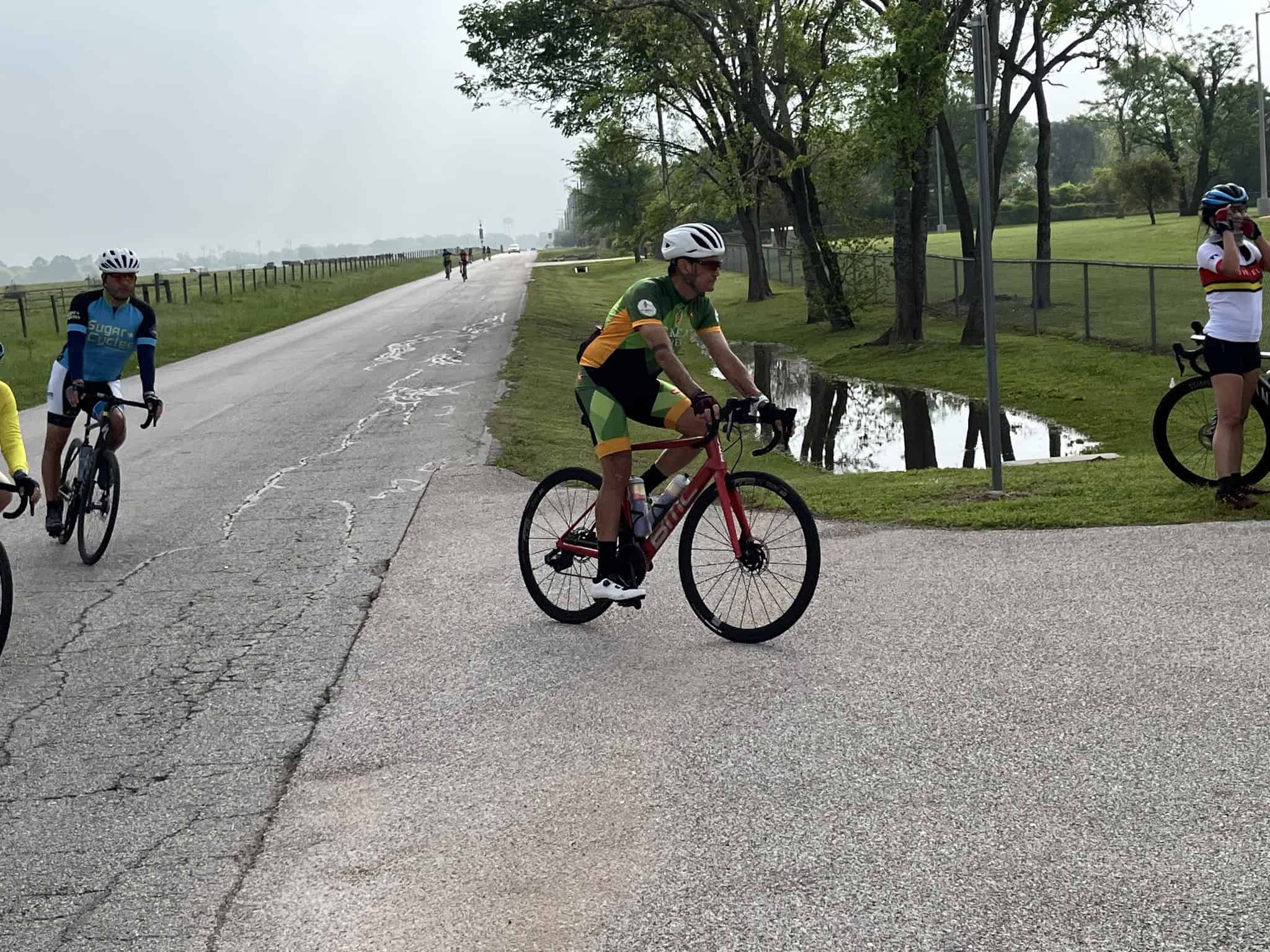 Cyclists riding bikes outdoors in a scenic park setting with trees and a small pond.