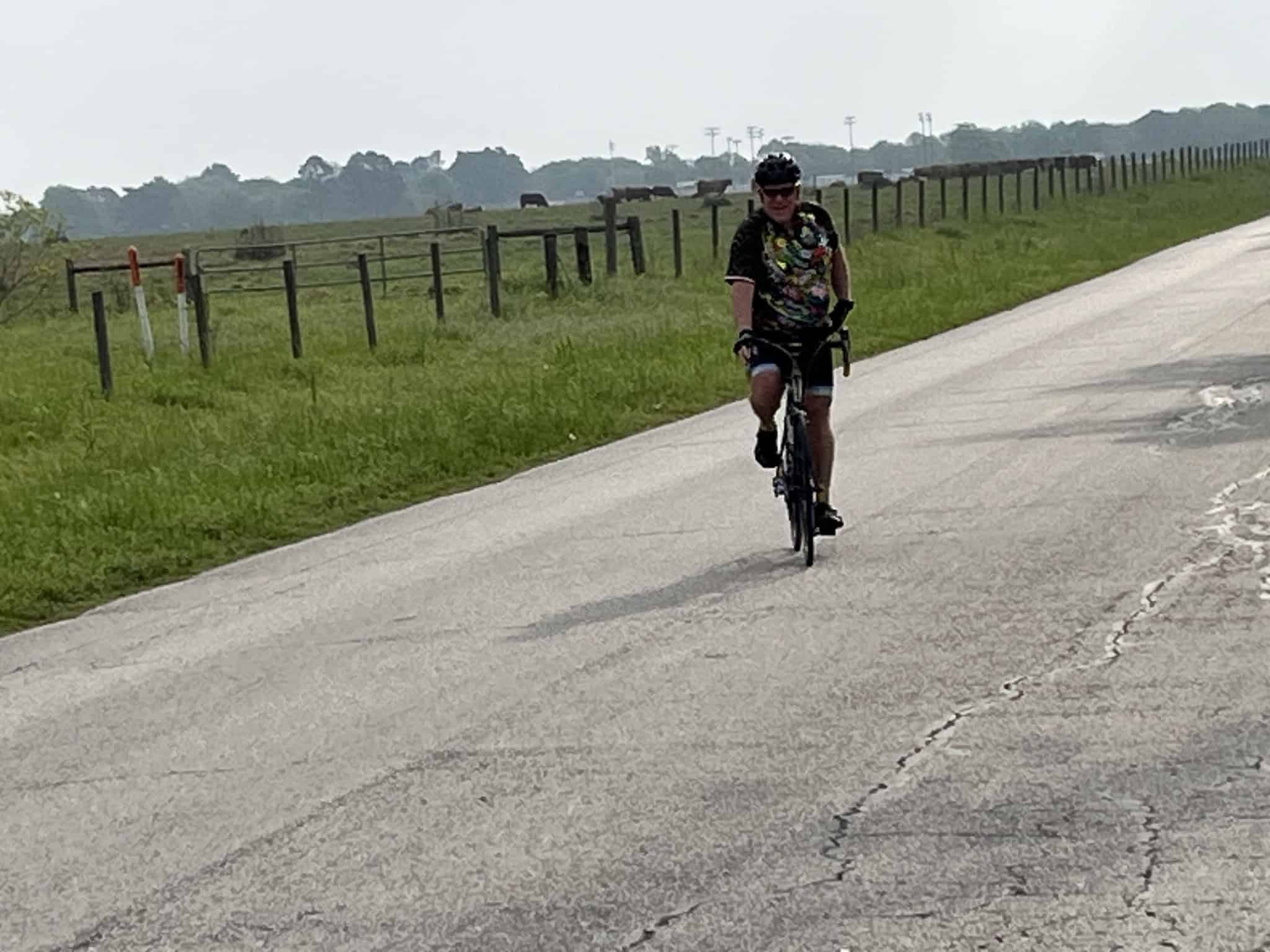 Bright woman riding a bicycle on country road with green fields and fences, sunny day - Bluebonnet Express.