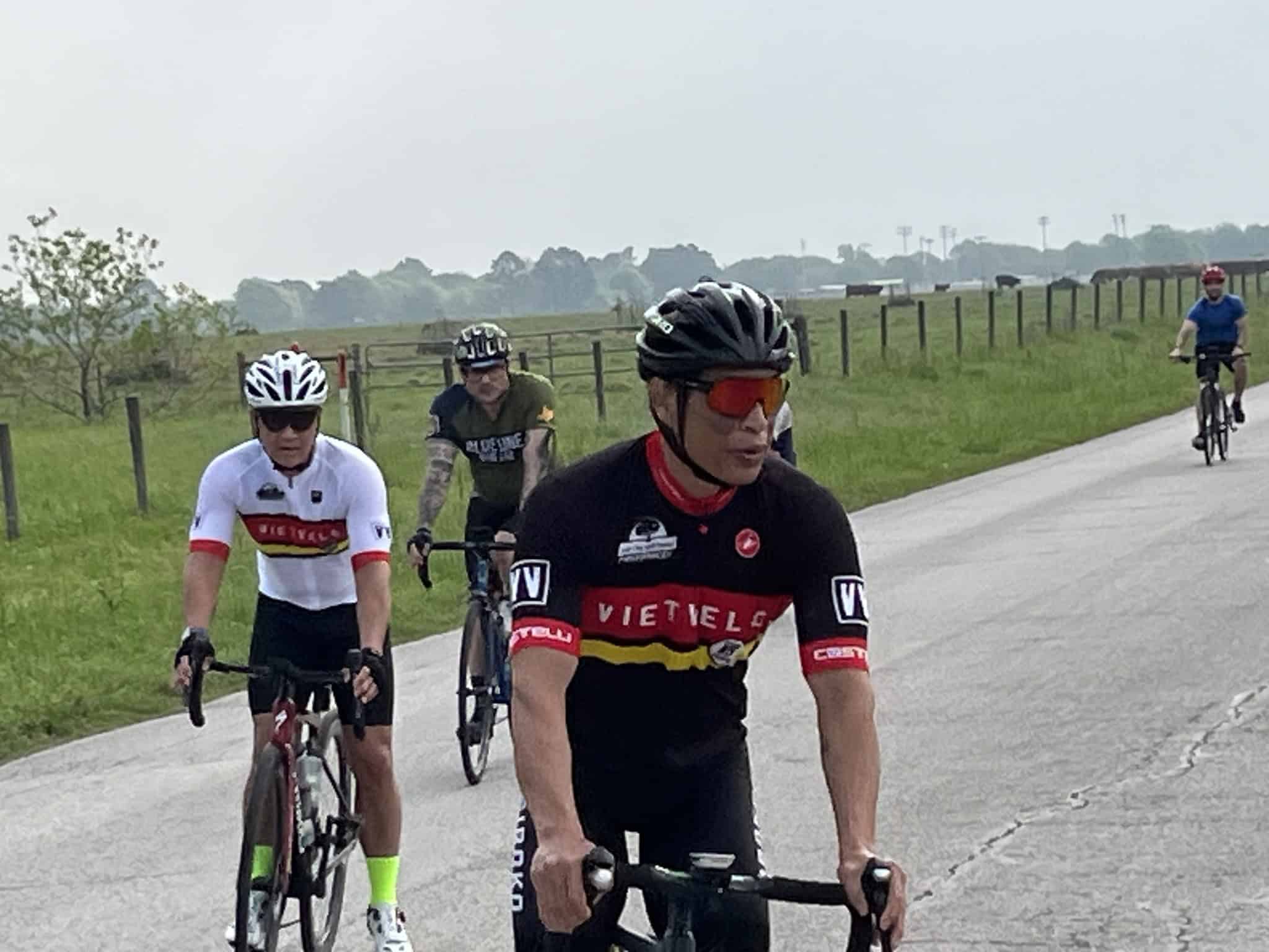 Riders cycling on open countryside road, representing Bluebonnet Express bike race, with scenic fields and wind turbines in background.