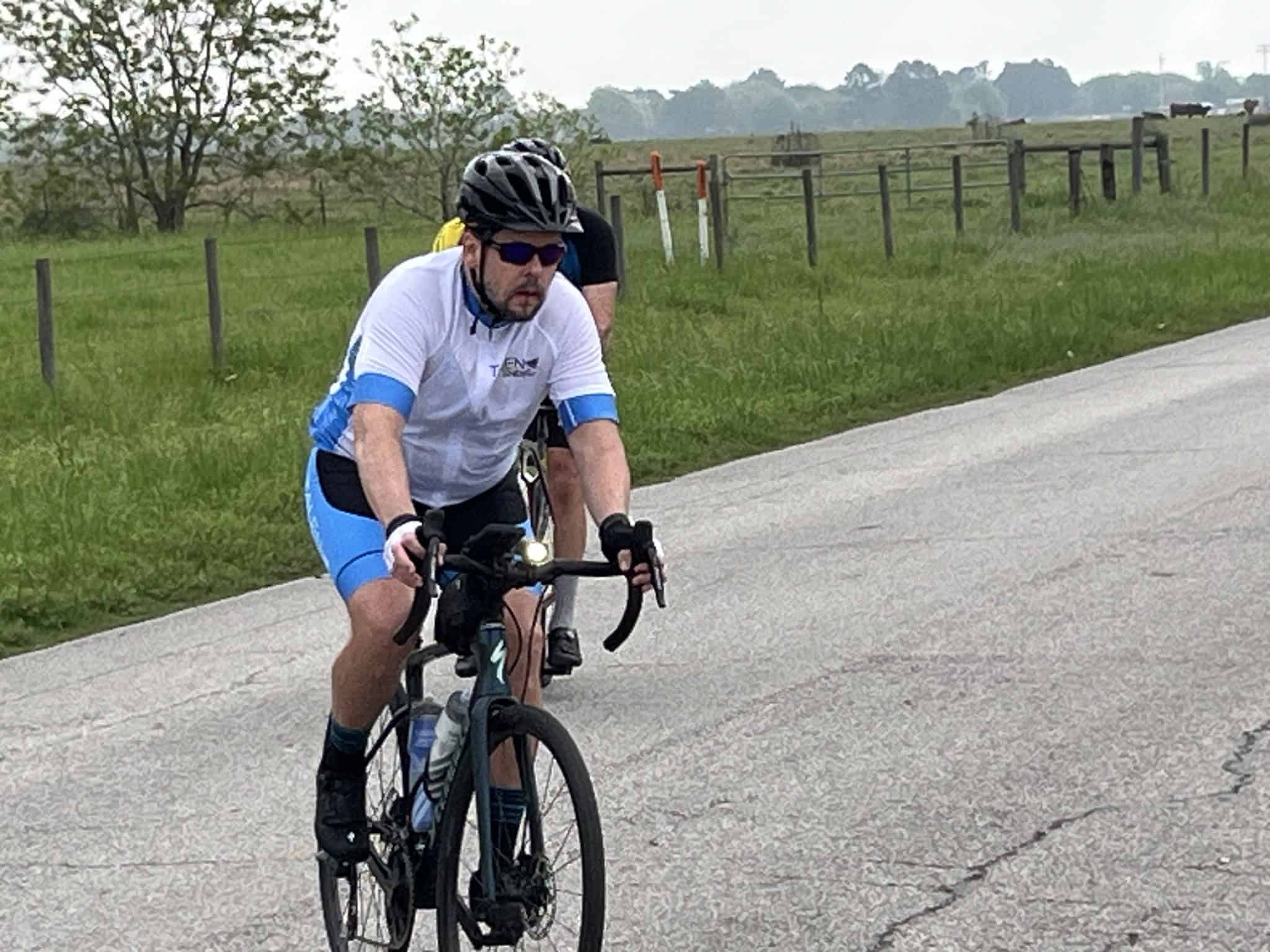 Cyclist riding on a rural road during daytime with green fields and fences in the background.