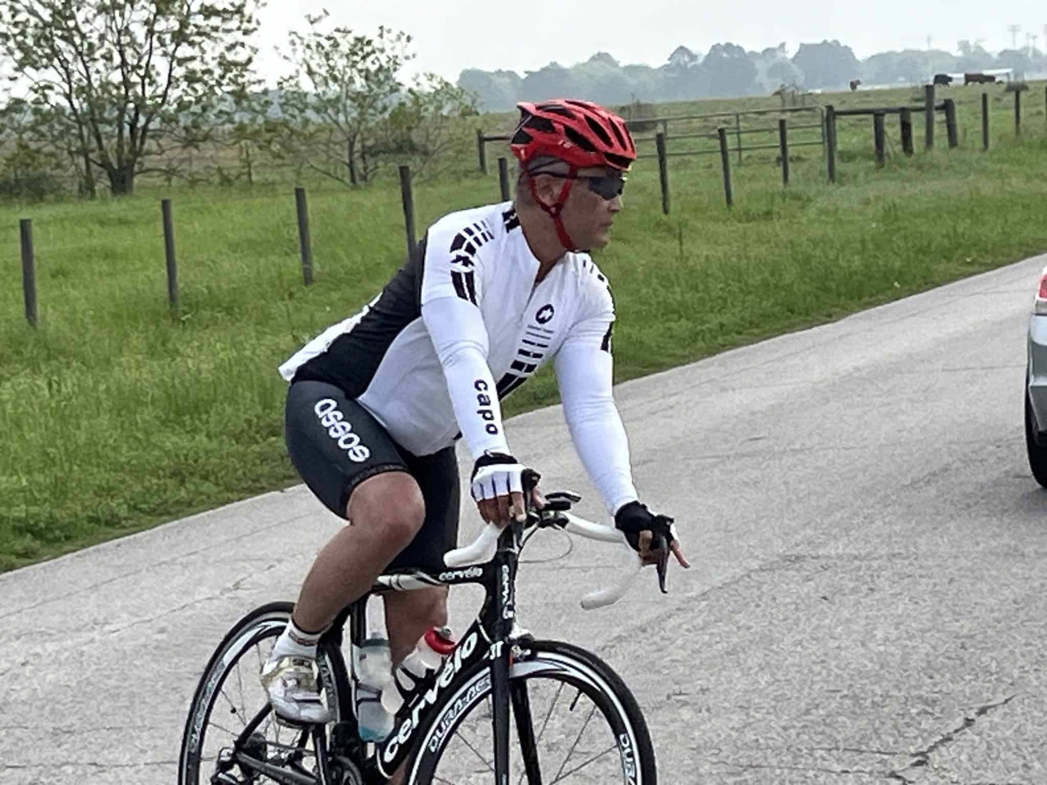 Velo cyclist wearing Bluebonnet Express cycling jersey and helmet riding on rural road.