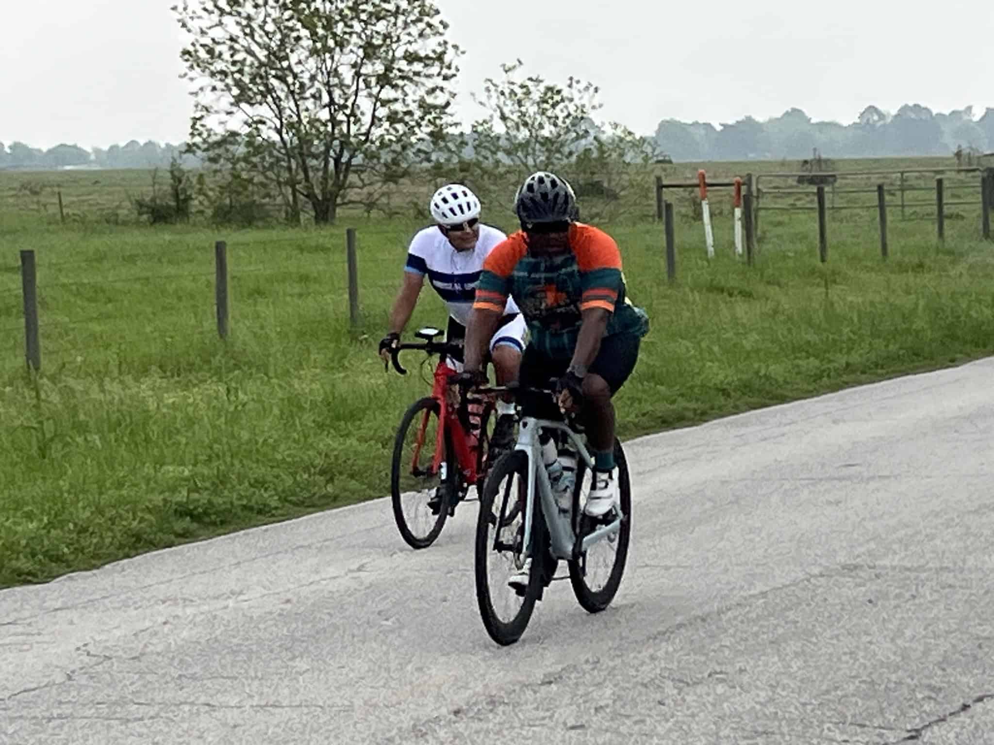 Riders cycling on rural road surrounded by green fields and fencing, enjoying outdoor cycling adventure.