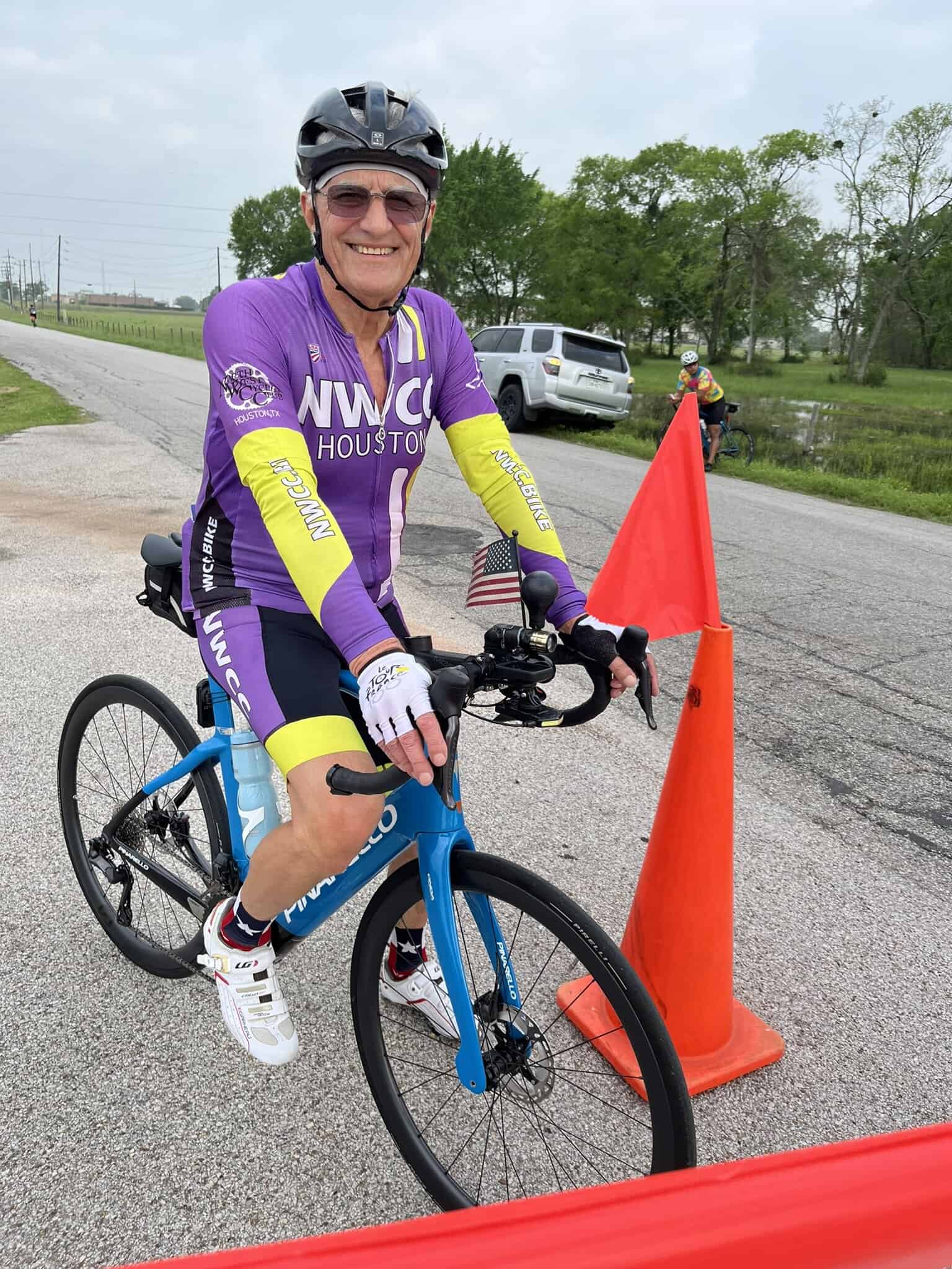 A man cycling at Bluebonnet Express event, smiling, wearing purple NWCC Houston cycling jersey, with orange cones and a scenic green background.