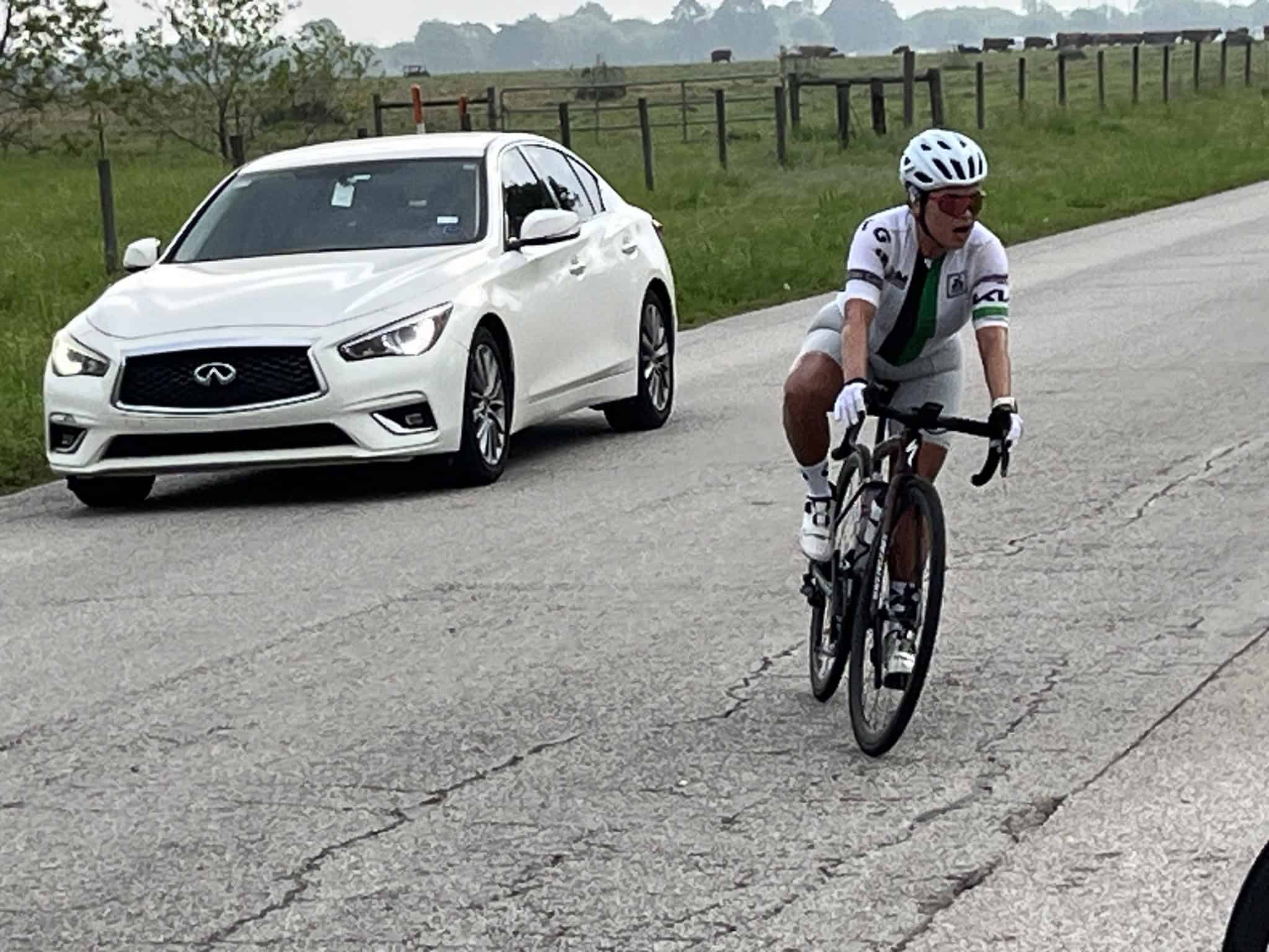 Cyclist riding on rural road with a white Infiniti car nearby, outdoor cycling event, scenic countryside backdrop.