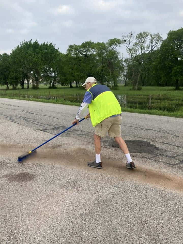 Loose asphalt patch repair on rural road using broom, road maintenance work, outdoor construction scene.