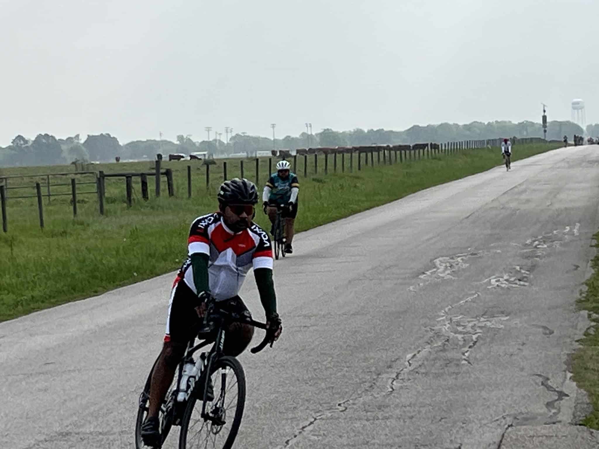 Riders cycling on open rural road with green fields and distant water tower in the background.