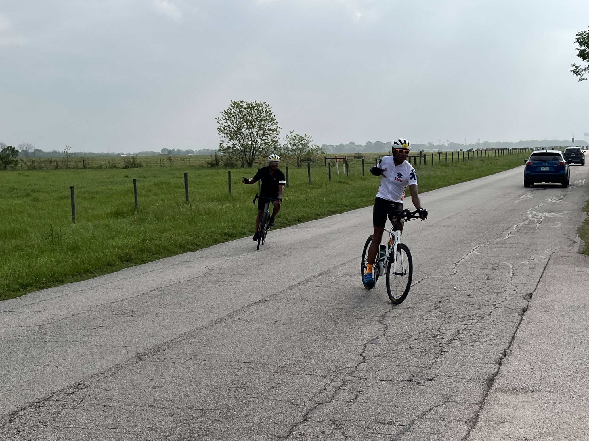 Cyclists riding on a rural scenic road with green fields and cars in the background.