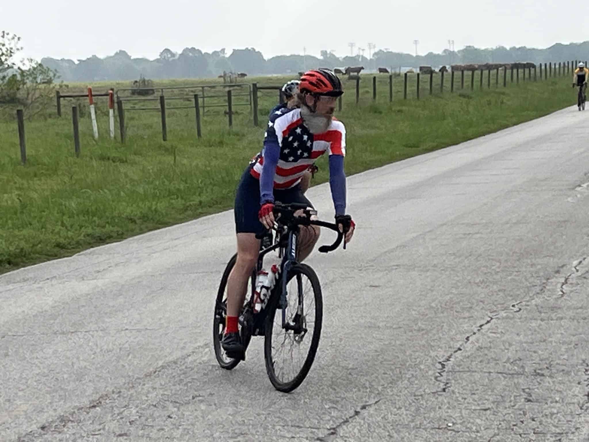 American flag-themed cycling jersey, road bike, outdoors, scenic rural route, cycling event, cyclist, Bike Ride, outdoor sports, cycling activity, fitness, adventure.