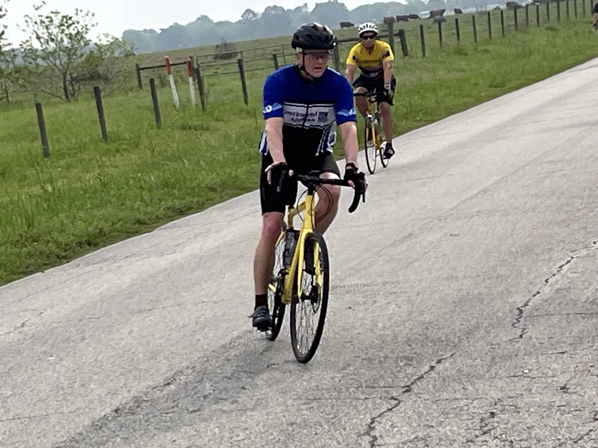Riders cycling on a rural road with green fields, promoting outdoor biking adventures with Bluebonnet Express.