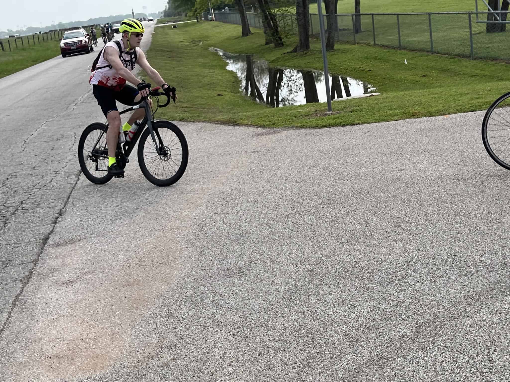 Cyclist riding a mountain bike on a paved trail near a grassy area with trees and a small waterway.