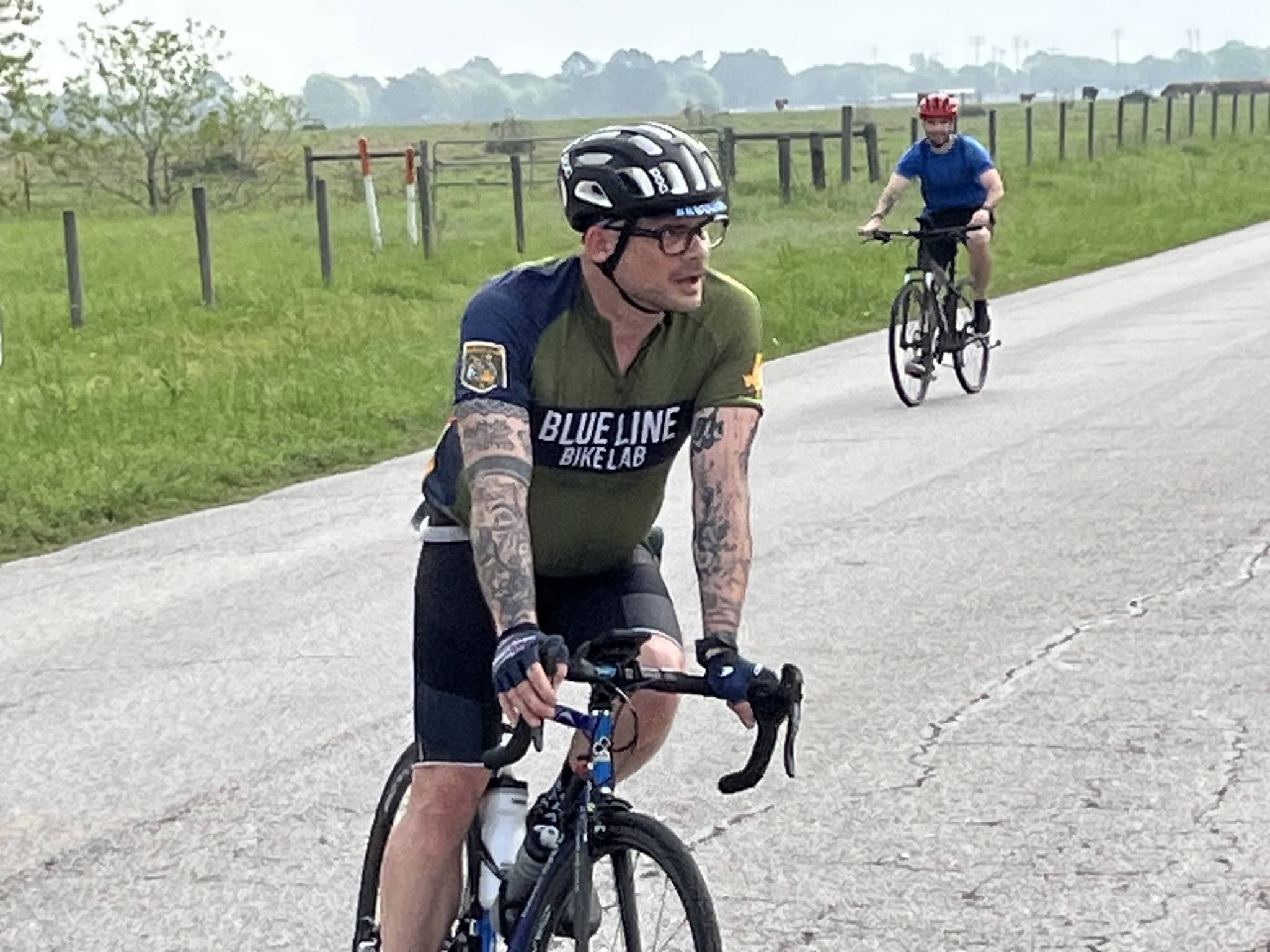 Tattooed cyclist wearing Bluebonnet Express jersey on a rural road during daytime.