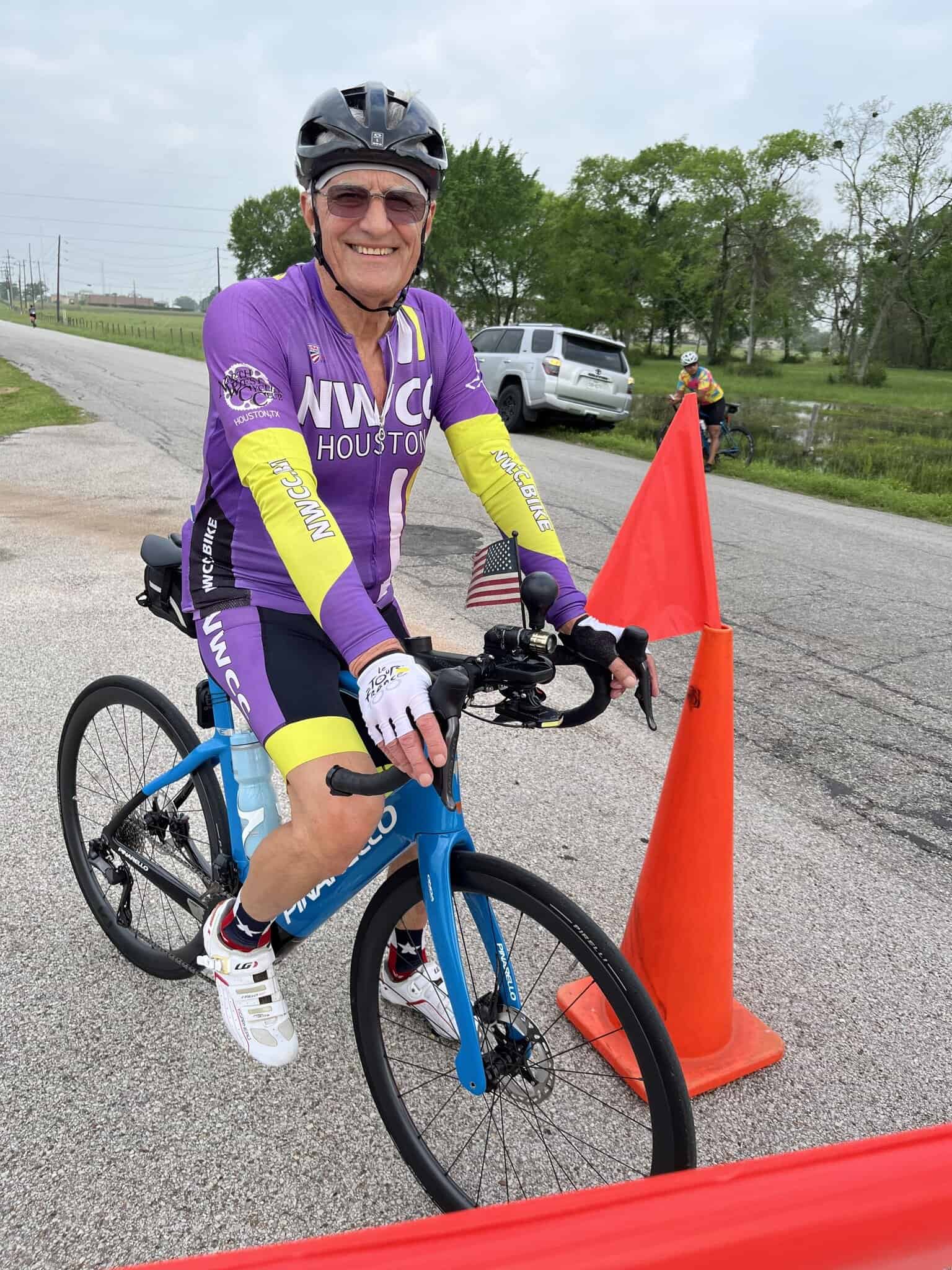 Vivid image of an older man smiling on a blue racing bike at the Bluebonnet Express cycling event. He wears a purple and yellow NWBC Houston jersey.