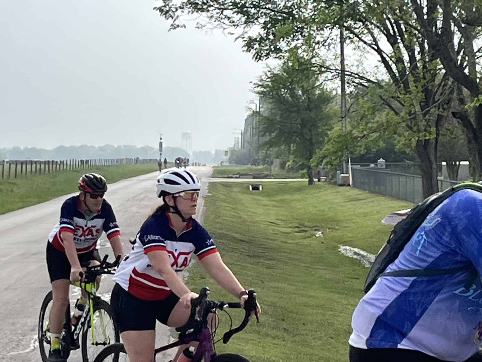 Cyclists riding on a scenic outdoor bike trail in daylight with green trees and open fields.