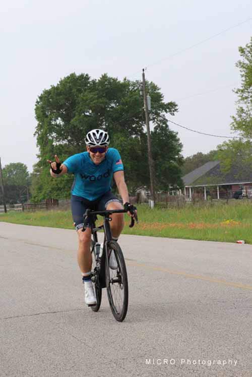Vegan cyclist riding on rural road during daytime, smiling and making peace sign, promoting Bluebonnet Express cycling event.