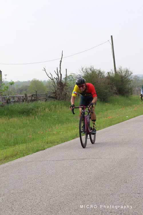 - A cyclist wearing a red and yellow jersey riding on a rural road during daytime.