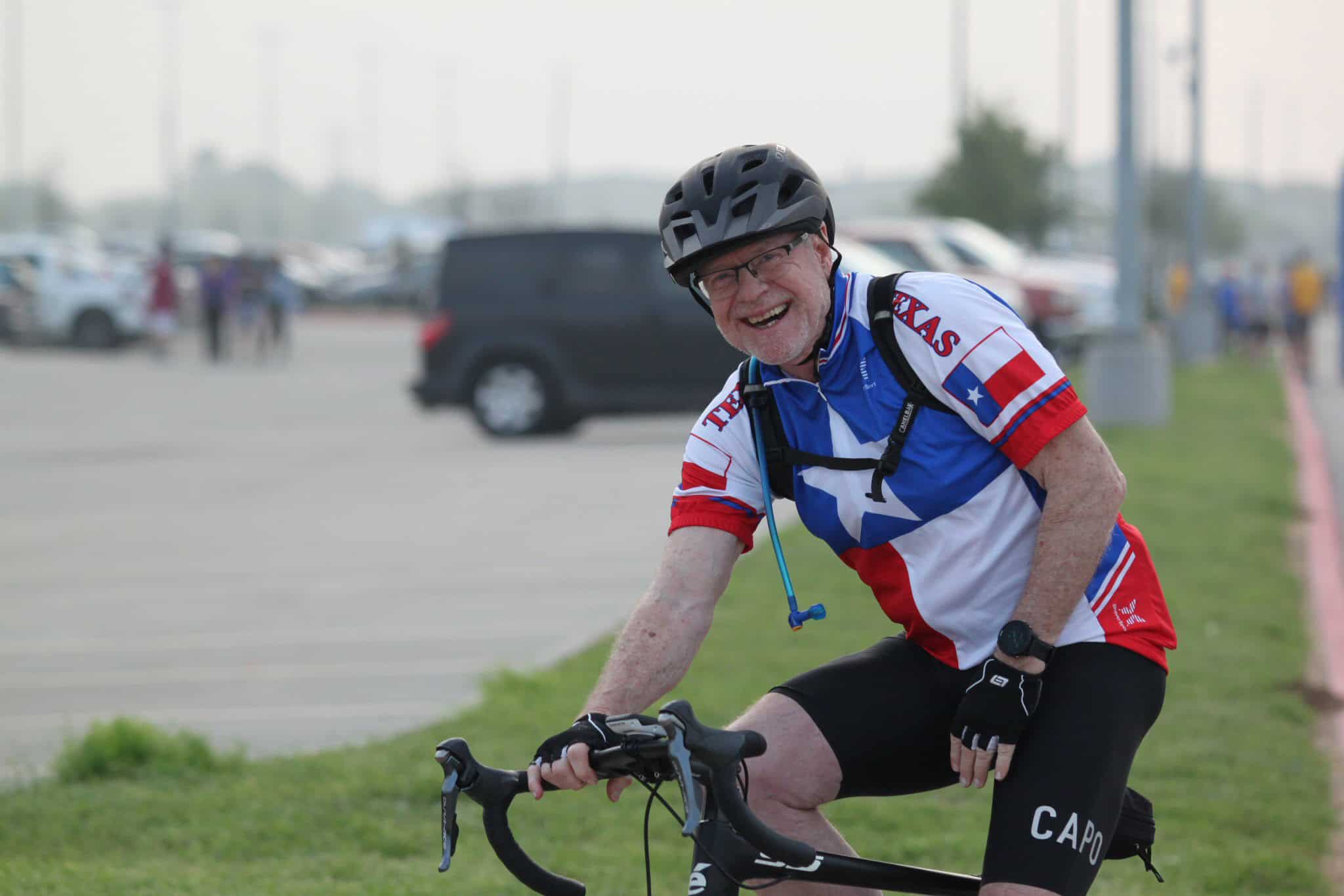 Bright smiling cyclist wearing Texas-themed jersey and helmet at Bluebonnet Express event at stadium parking lot.