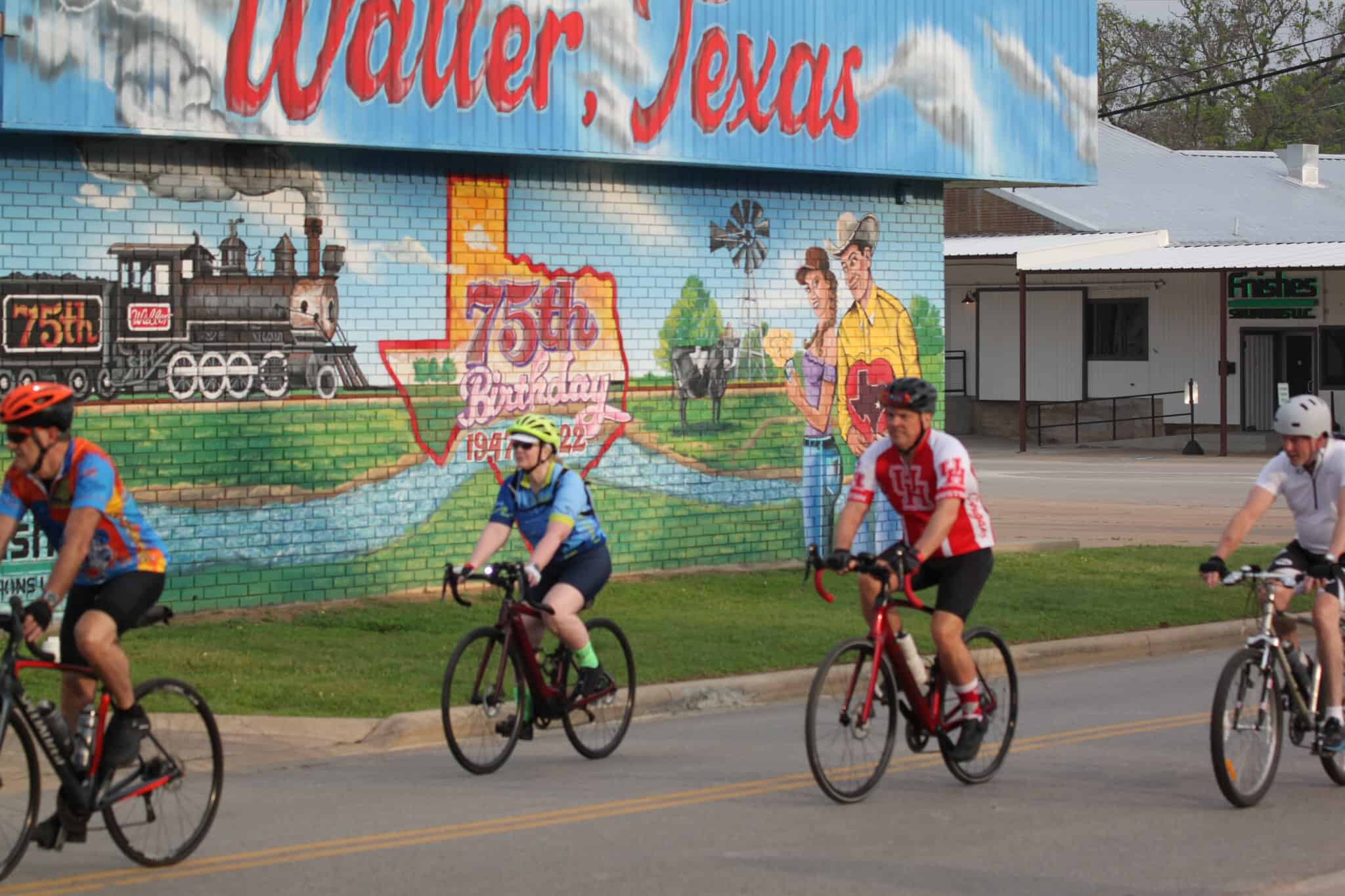 Colorful mural celebrating Water, Texas 75th birthday behind cyclists on a bike ride route.