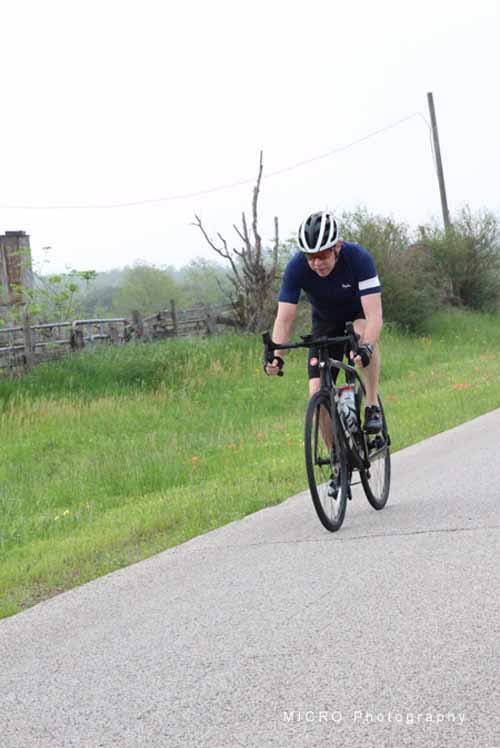 Velo cyclist riding on a scenic rural road with greenery and open sky in the background.
