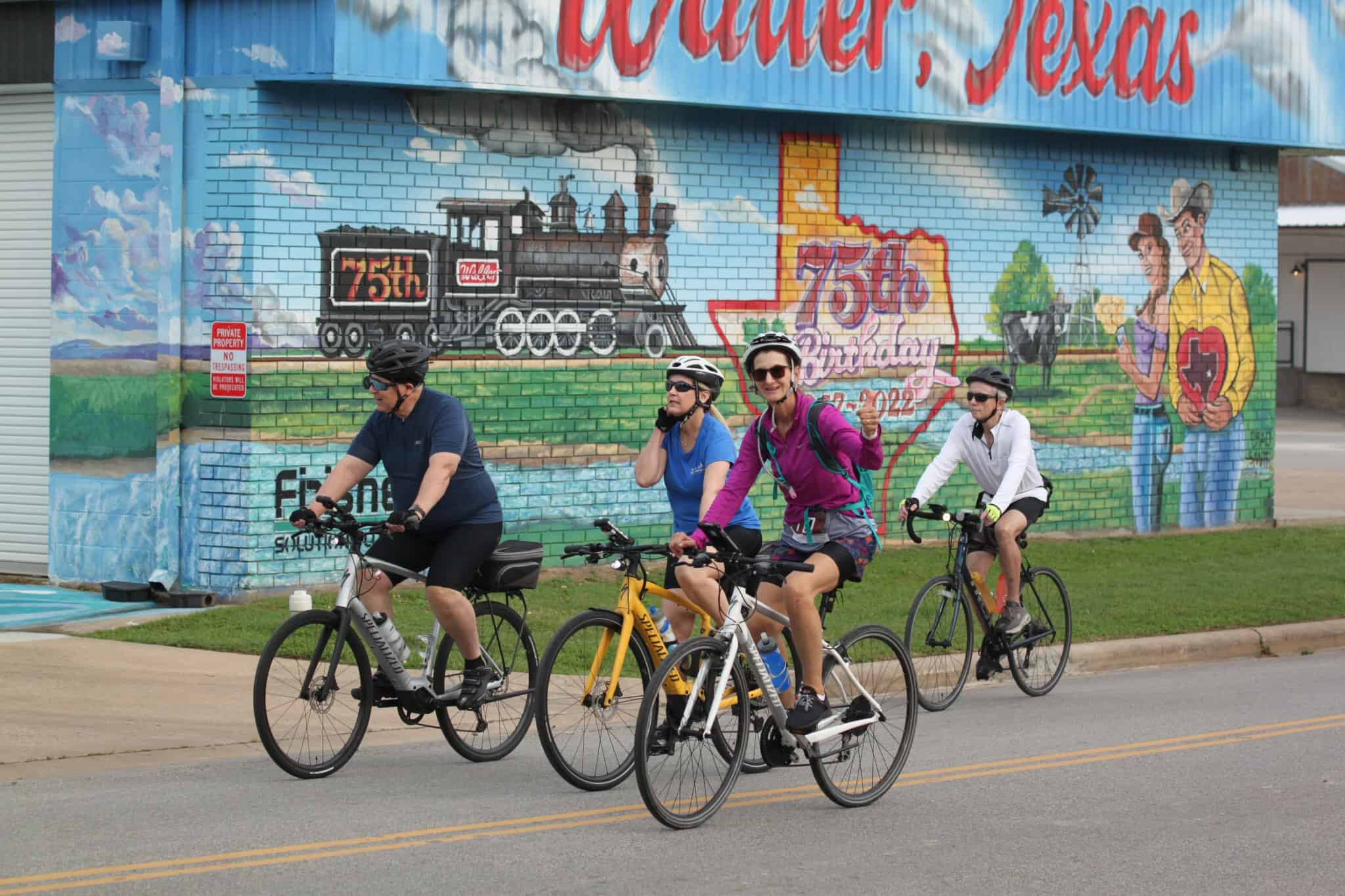 Bright group of cyclists riding past a colorful mural celebration of Bluebonnet Express 75th birthday in Waxahachie, Texas.