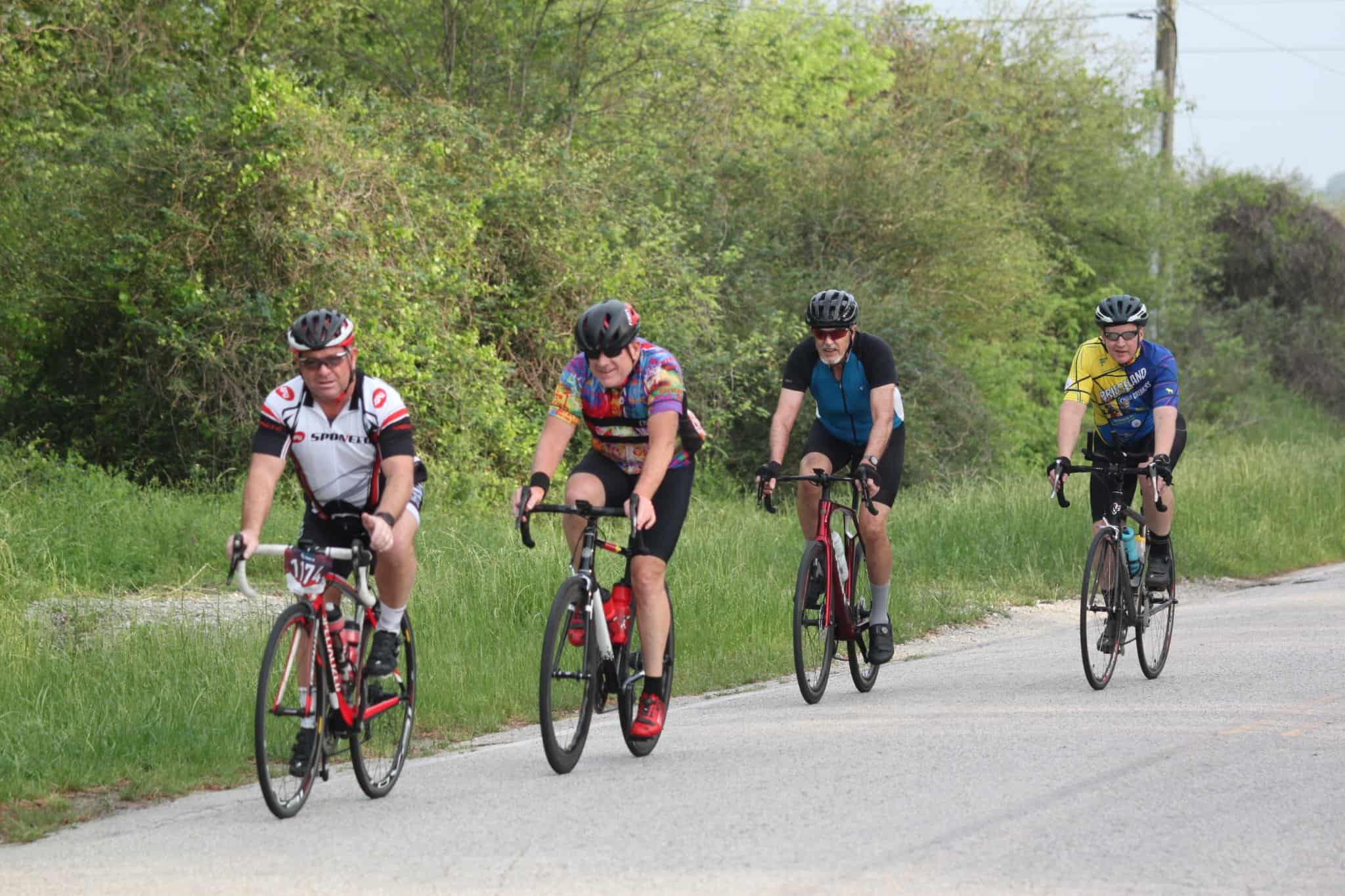 Brightly colored cyclists riding along scenic rural road with lush greenery and trees, promoting outdoor fitness and biking events.